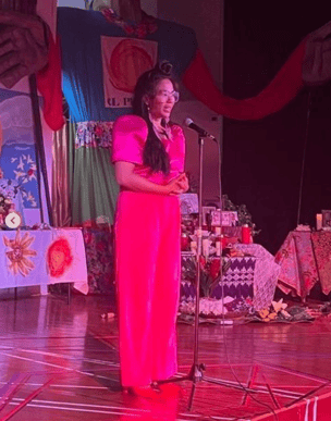 Jana Lynne Umipig wears a pink blouse and pants and stands in front of a microphone on the stage of El Puente.