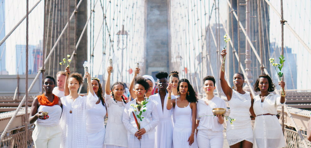 The healers of Harriet’s Apothecary wearing all white, stand together lifting up flowers and candles before the backdrop of the Brooklyn Bridge.