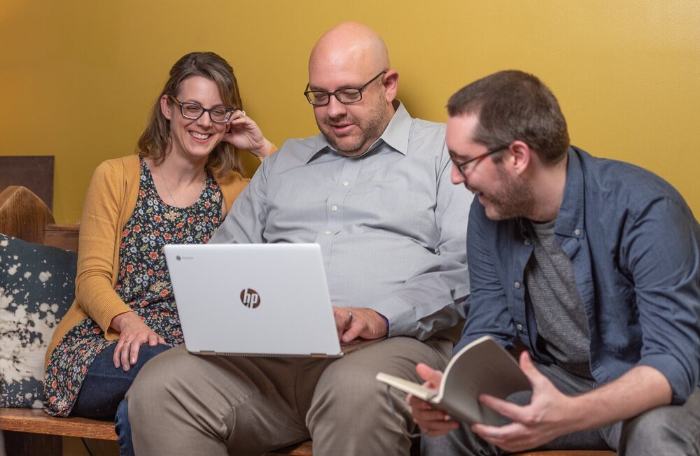 A woman and two men, named Lori, Chris, and Jarred, sit on a couch in front of a yellow wall, smiling and looking at the same laptop being held by the man sitting in the middle.